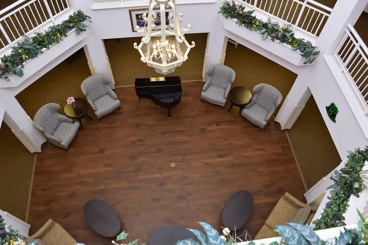 View from above of a senior living facility common area with four patterned armchairs arranged around two small round tables, a black grand piano in the center, a chandelier hanging from the ceiling, and decorative greenery along the railings of the upper floor.