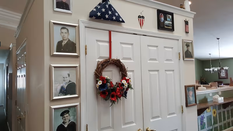 Interior view of a hallway in a senior living facility with white double doors decorated with a floral wreath. Surrounding the doors are framed black-and-white and color photographs of military personnel, a folded American flag in a triangular display case, and patriotic decorations. The adjacent room has green walls and a dining area with a chandelier and tables.