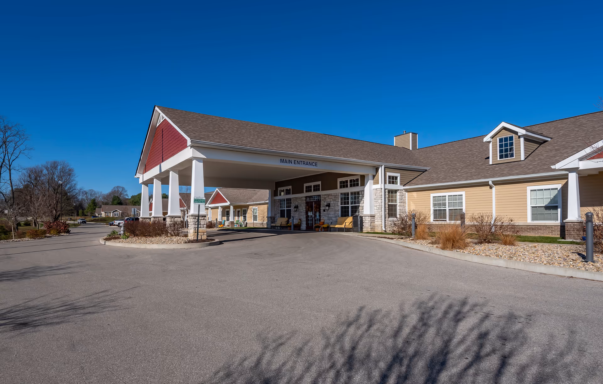 Exterior view of the main entrance of a senior living facility with a covered driveway, beige siding, stone accents, and a clear blue sky.