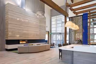 Spacious modern interior of a senior living facility featuring a large beige sectional sofa facing a long horizontal fireplace set in a tall wall with a light wood finish. The room has high ceilings with large windows allowing natural light, contemporary pendant lights hanging over a white kitchen island with bar stools, and wooden beams overhead.