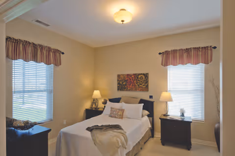 A cozy bedroom with a neatly made bed featuring white linens and decorative pillows. The room has two windows with striped valances, two bedside tables each with a lamp, a piece of abstract artwork above the bed, and a dresser with decorative items on it.