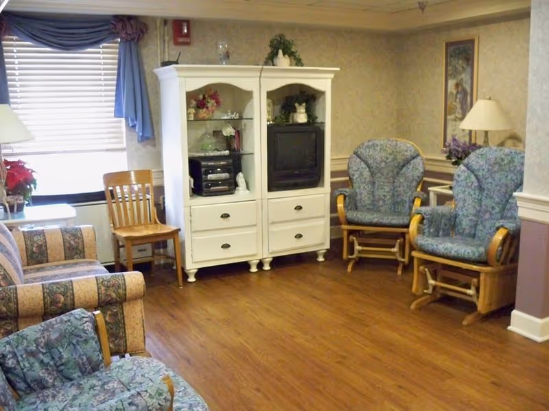 Cozy senior living room with upholstered chairs, a white entertainment cabinet holding a TV and stereo, and wood flooring.