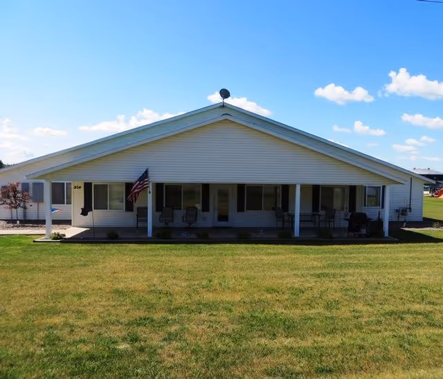 Single-story white building with a covered front porch, chairs and an American flag facing a grassy lawn under a blue sky.