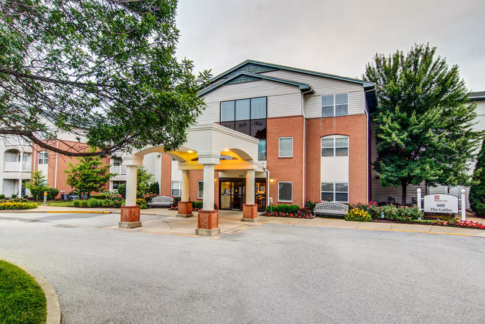 Front entrance of a three-story senior living building with a covered portico, benches, and landscaped grounds.
