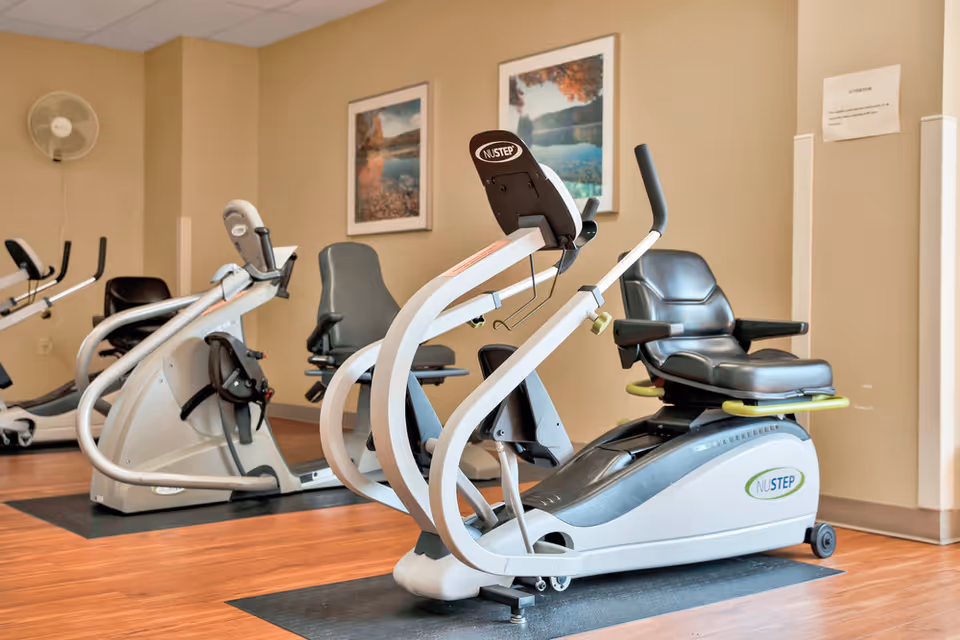 A small exercise room with two recumbent stationary bikes on black mats placed on a wooden floor. The walls are beige with two framed landscape pictures hanging. A wall-mounted fan is visible in the corner, and a white sign is posted on the wall.