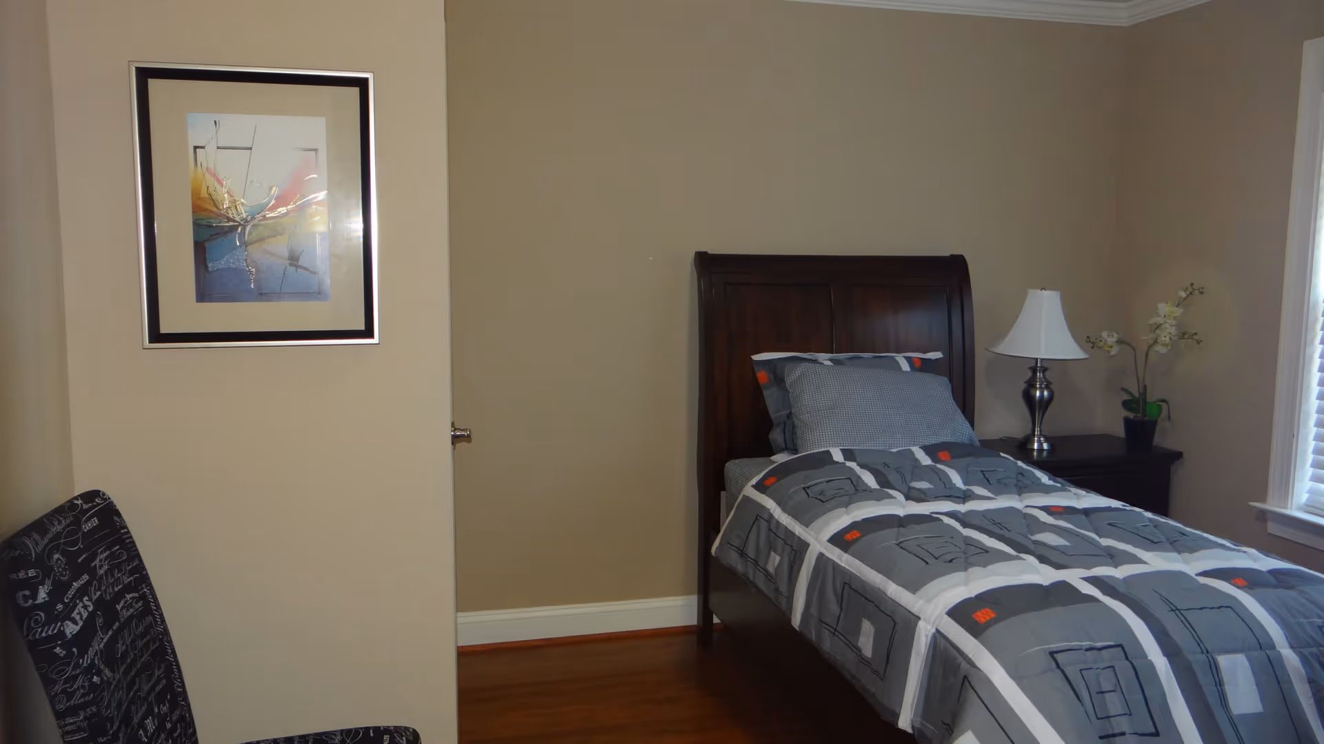 A small bedroom with a single wooden bed featuring a gray and white patterned comforter and pillows. Next to the bed is a dark wooden nightstand with a silver lamp and a small potted plant with white flowers. A window with white blinds is partially visible on the right side, and a framed abstract artwork hangs on the beige wall to the left. A chair with a black and white script pattern is partially visible in the foreground.