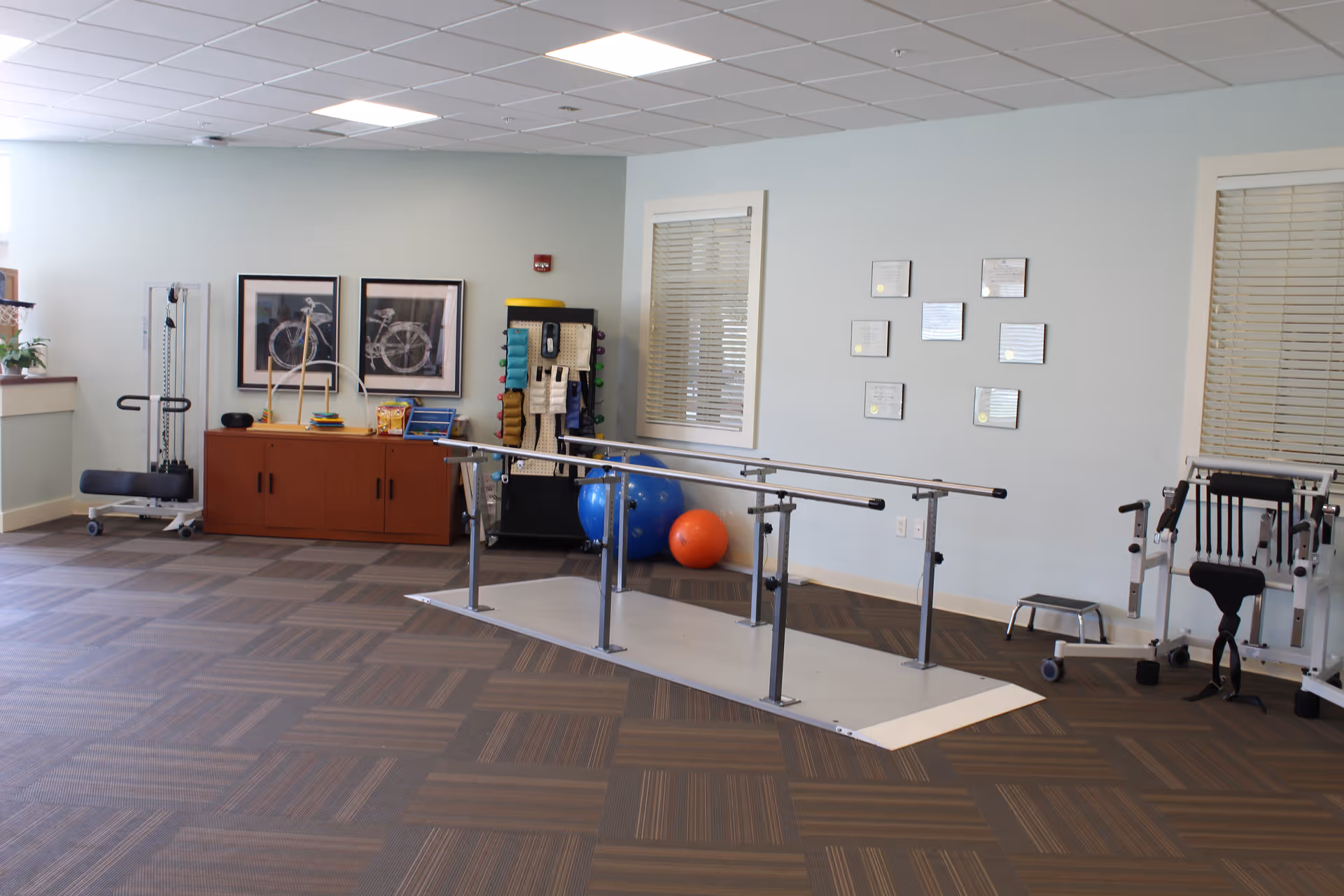 Therapy room with parallel walking bars, exercise balls, and other rehabilitation equipment against a pale blue wall.