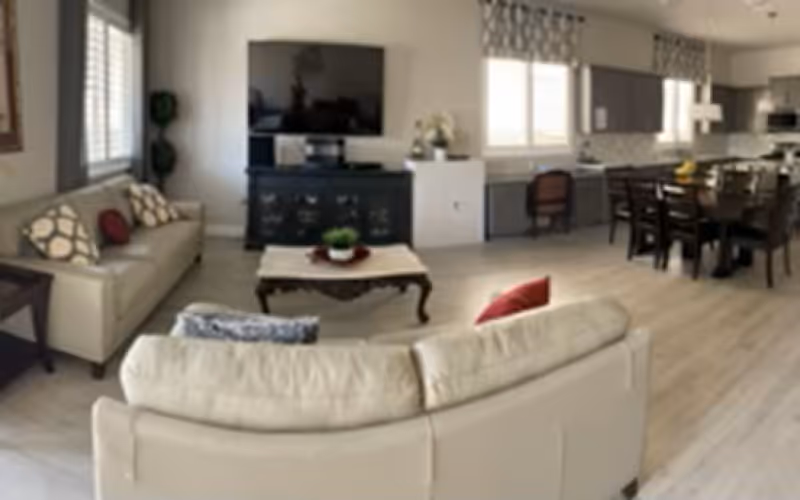 Open-plan living area with beige sofas around a coffee table facing a wall-mounted TV, with a dining table and kitchen visible in the background.