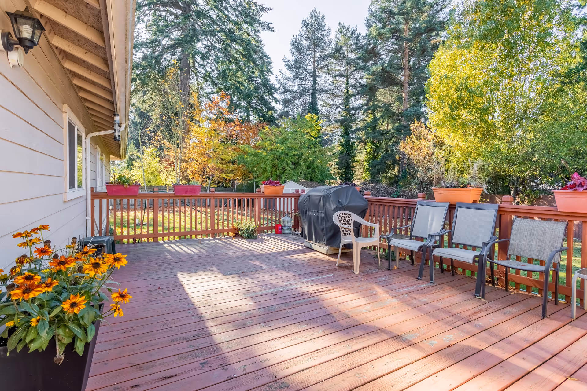 A spacious wooden deck attached to a house, featuring several chairs, a covered grill, and flower pots with colorful flowers. The deck overlooks a green yard with trees showing autumn foliage under a clear sky.