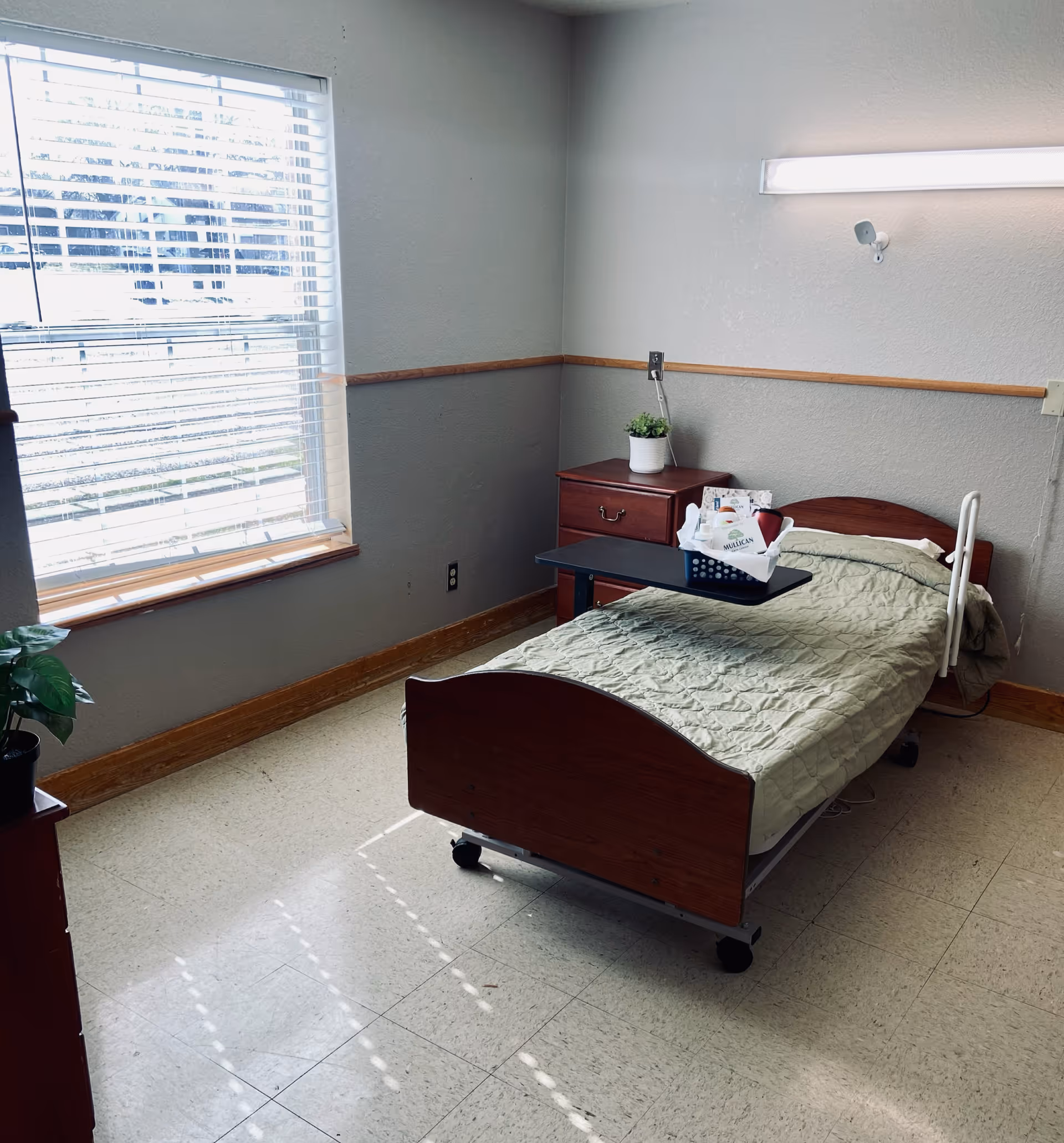 A simple, clean bedroom in a care facility with a single hospital-style bed covered with a green quilt. Next to the bed is a wooden nightstand with a small potted plant and a basket containing items labeled Mullican. There is a large window with blinds letting in natural light, and the walls are painted gray with a wooden trim.