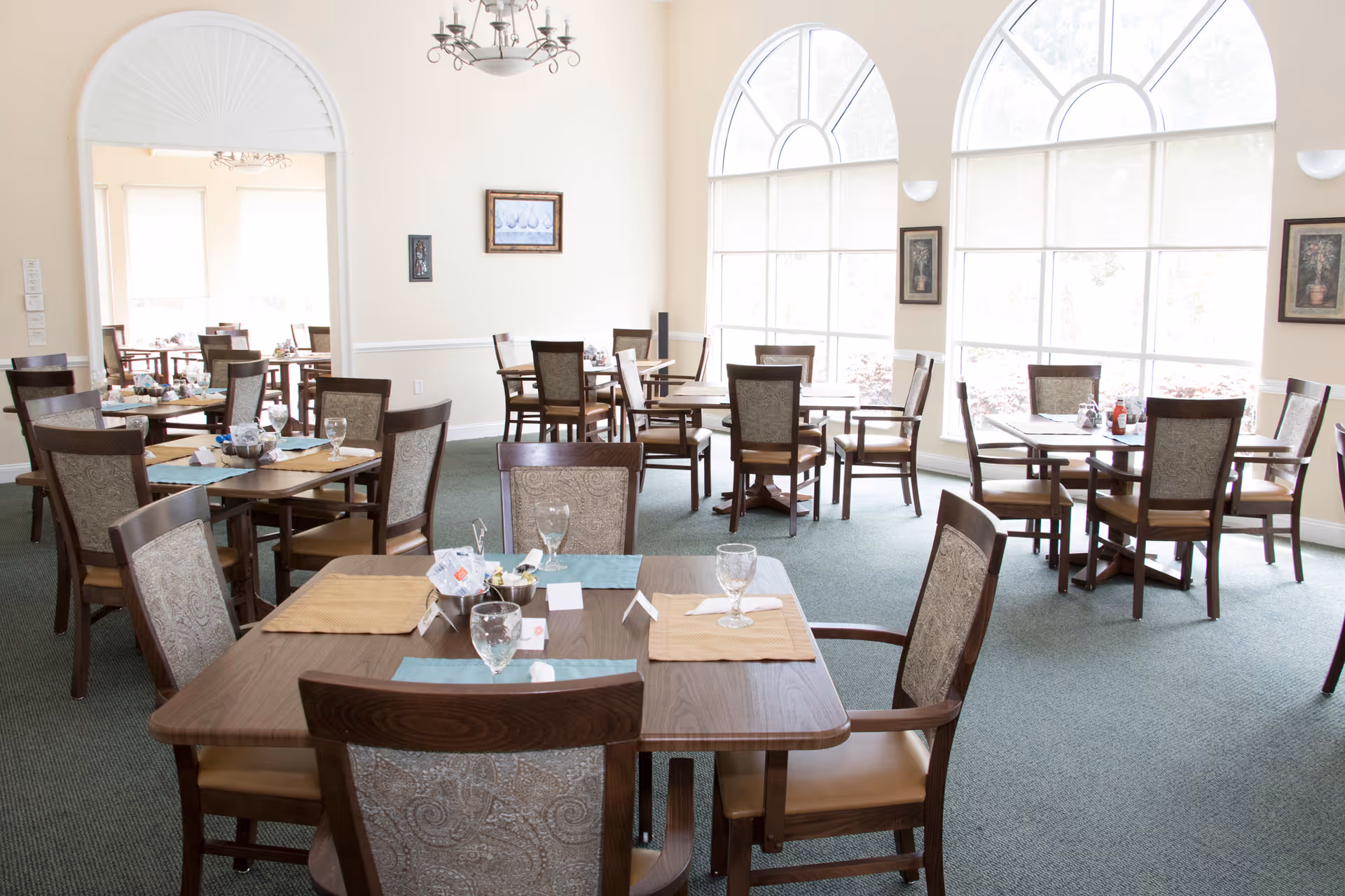 Sunlit dining room with multiple wooden tables and chairs set with placemats and glasses in front of large arched windows.