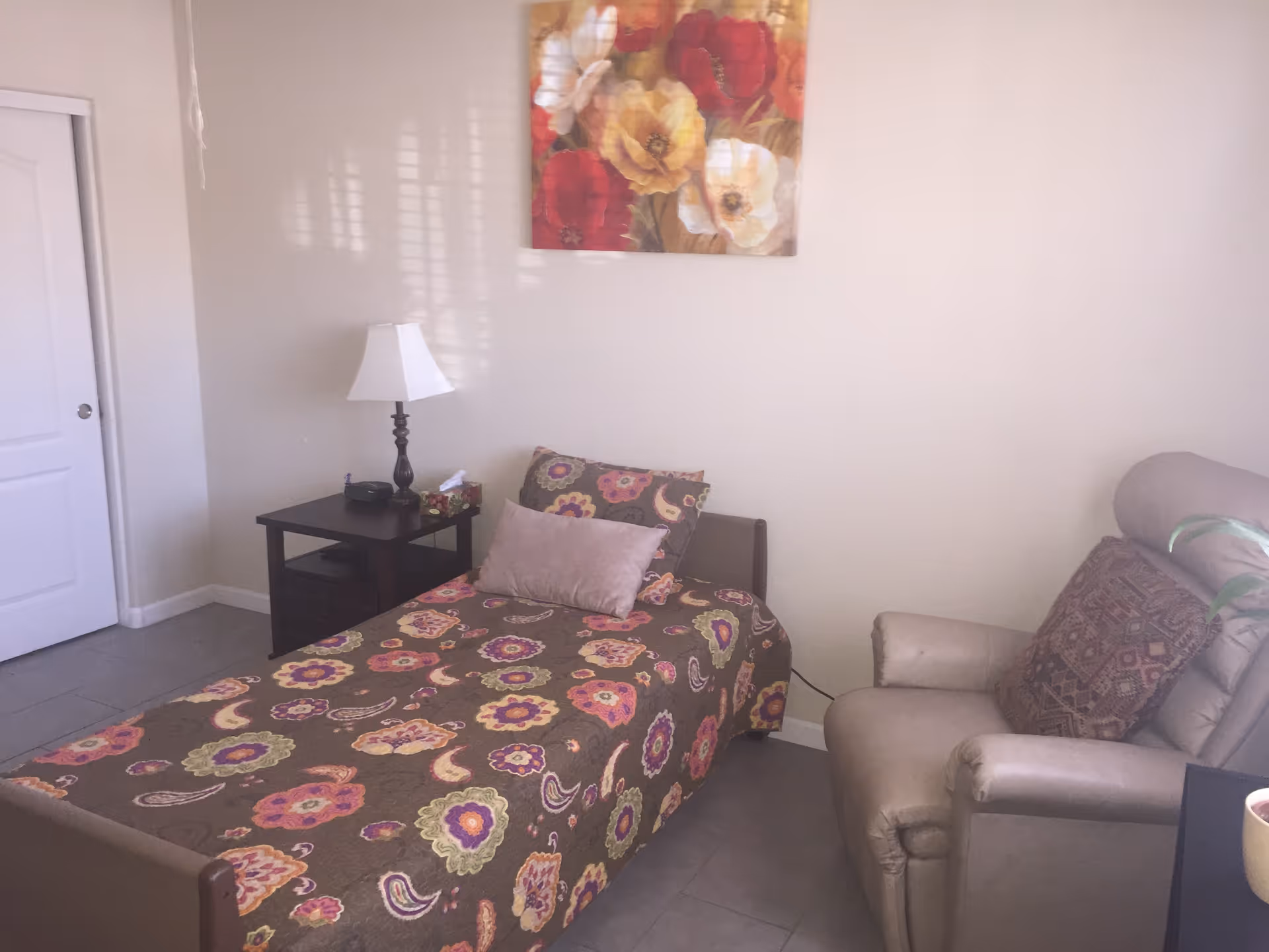A small bedroom with a single bed covered in a colorful floral and paisley patterned bedspread. There are two pillows on the bed, one plain and one matching the bedspread. Next to the bed is a dark wooden nightstand with a white lampshade, a tissue box, and a small black item. On the right side of the room is a beige recliner chair with a patterned cushion. A painting of large flowers in red, yellow, and white hangs on the wall above the bed. The room has light-colored walls and a closed white door on the left side.