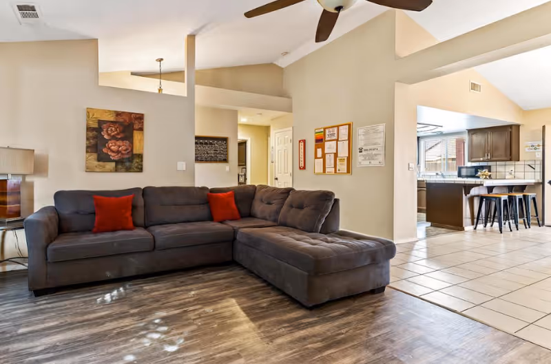 Open living room featuring a gray sectional sofa with red throw pillows, wood-look flooring, and a view into a tiled kitchen area.