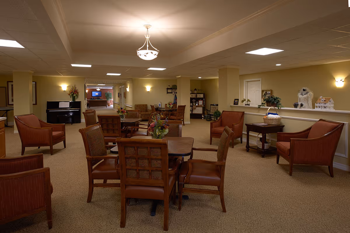 A spacious and warmly lit common area with multiple wooden chairs and tables arranged for seating. The room features beige walls and carpet, wall sconces, a piano on the left side, and decorative plants and flowers placed on tables and shelves. There is a small table with a basket and a dollhouse on the right side, and a hallway leading to another room in the background.