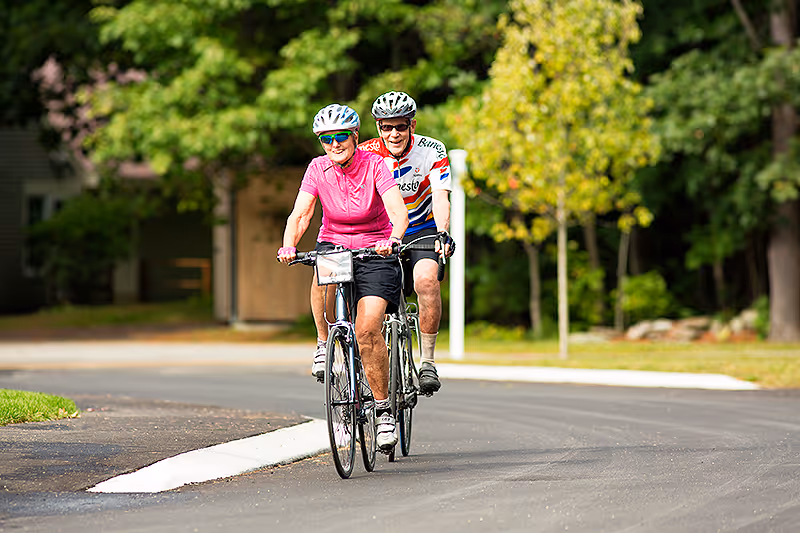 Two elderly people wearing helmets and cycling on a paved road surrounded by green trees and grass, enjoying an outdoor bike ride.
