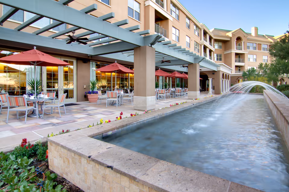 Outdoor patio area at Vi at Silverstone with tables and chairs under red umbrellas next to a long rectangular water feature with a fountain, adjacent to a multi-story building.