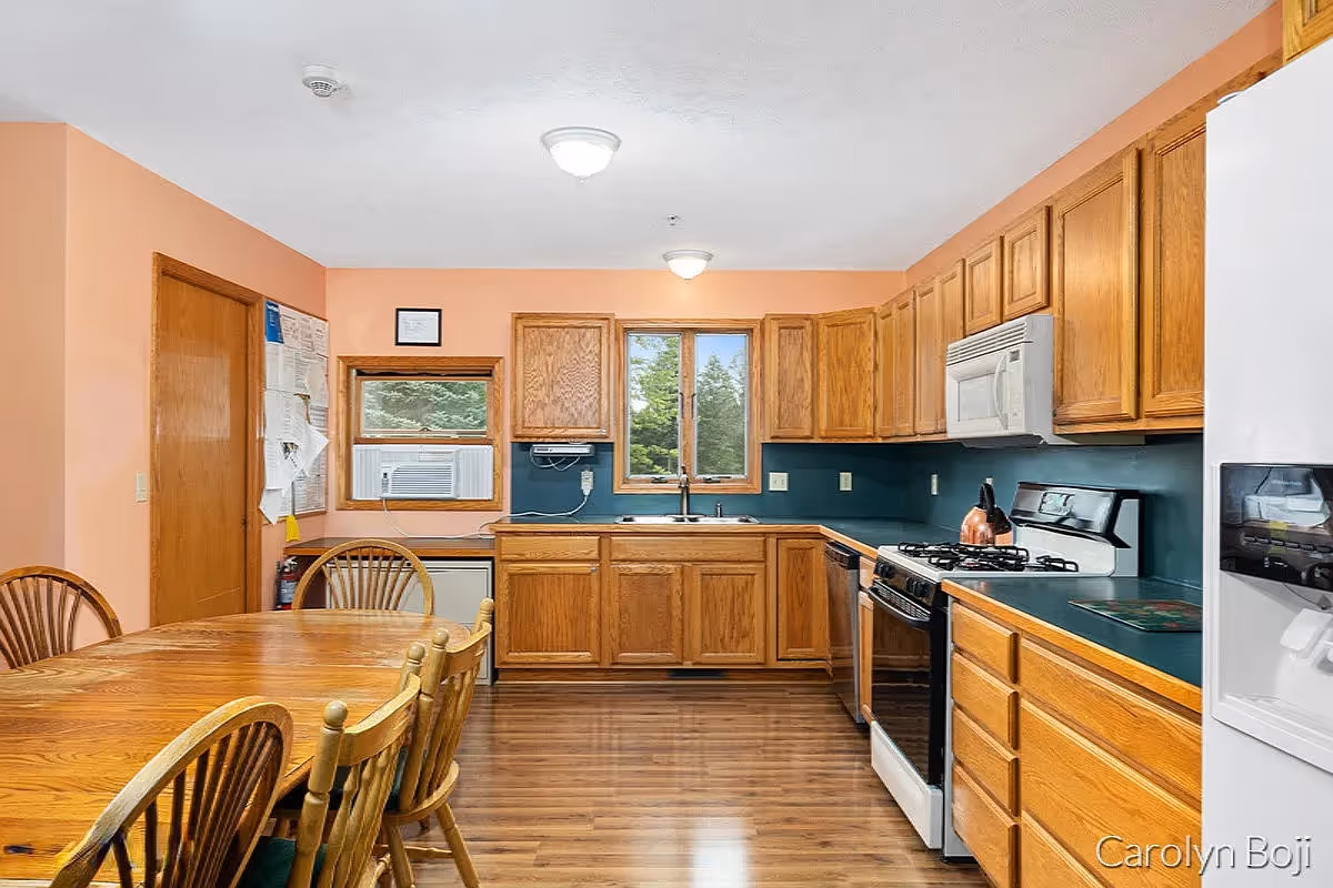 A kitchen with wooden cabinets and a wooden dining table with six chairs. The kitchen features a gas stove with a kettle, a microwave, a dishwasher, a refrigerator with an ice dispenser, and a window with a view of trees. The walls are painted peach, and the floor is wooden.