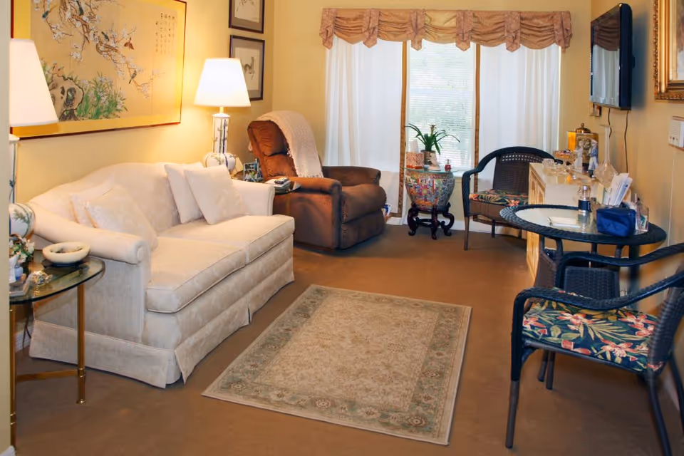 A cozy living room with a beige sofa, a brown recliner chair, a glass side table with a lamp, a patterned area rug, and a small round table with two floral cushioned chairs. The room has light yellow walls adorned with framed artwork and a large window with white curtains and a valance.