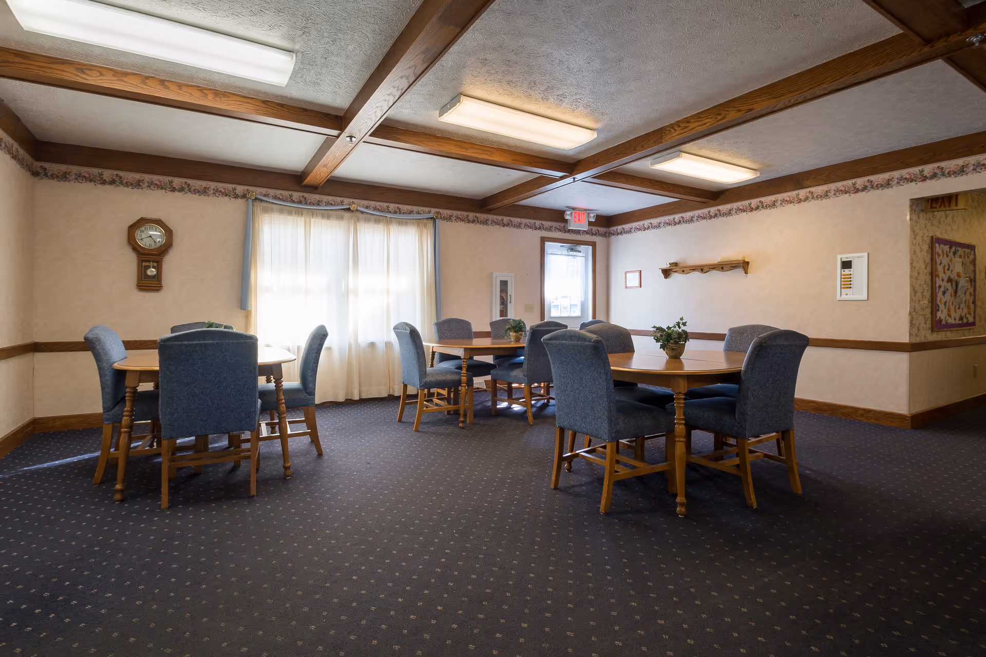 A quiet dining room with two wooden tables, each surrounded by six blue upholstered chairs. The room has beige walls with floral wallpaper border near the ceiling, a carpeted floor, and wooden ceiling beams. There is a window with sheer curtains letting in natural light, a wall clock, and some small potted plants on the tables.