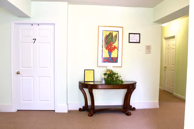 A hallway in an elder care facility with a white door labeled number 7 on the left, a wooden console table with a plant and framed documents on it against a white wall, and a colorful framed artwork hanging above the table. Another door is visible down the hallway on the right.