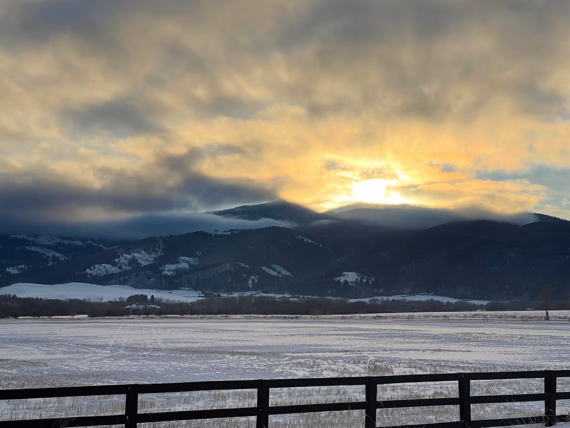 A scenic view of snow-covered fields with a black wooden fence in the foreground, mountains in the background partially covered by clouds, and a bright sun setting or rising behind the mountains casting a warm glow in the cloudy sky.