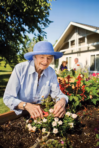 An elderly woman wearing a blue hat and light blue shirt is gardening, tending to flowers in a garden bed outside a house on a sunny day.