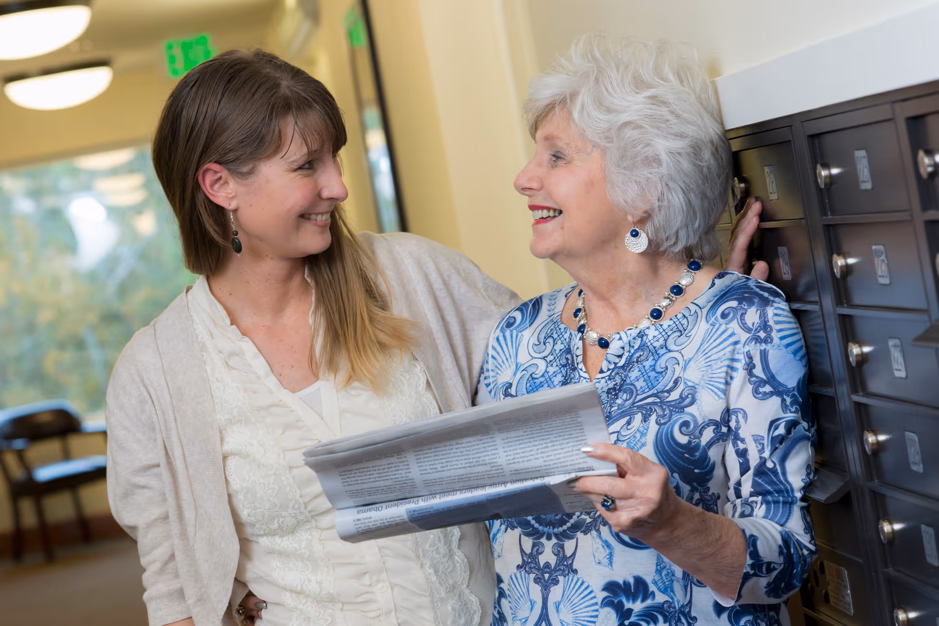 An elderly woman and a younger woman smiling and talking while standing next to a row of mailboxes in a hallway. The elderly woman is holding a newspaper, and both appear happy and engaged in conversation.