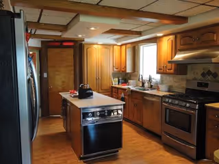 Interior view of a kitchen with wooden cabinets, a central island with a dishwasher, a stainless steel refrigerator, a stove with an oven, a window above the sink, and various kitchen items on the counters.