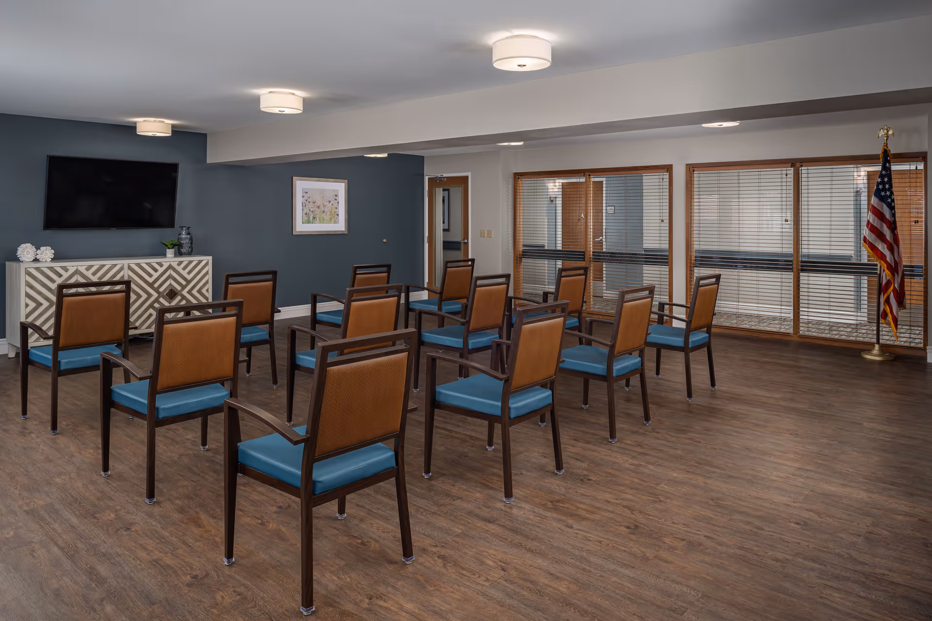 A senior living facility common room with multiple wooden chairs featuring blue cushions arranged in rows facing a wall-mounted flat screen TV. The room has wood flooring, a dark blue accent wall with a framed picture, a patterned sideboard, and large windows with wooden blinds. An American flag stands in the corner near the windows.