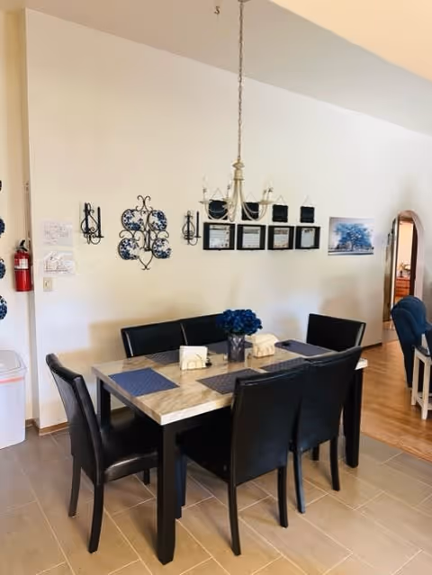A dining area with a rectangular marble-top table surrounded by six black chairs. The table is set with blue placemats and a centerpiece of blue flowers. The wall behind the table features decorative blue and black wall art, a chandelier hanging above the table, and framed pictures. A fire extinguisher is mounted on the left wall, and an arched doorway leads to another room with wooden flooring.