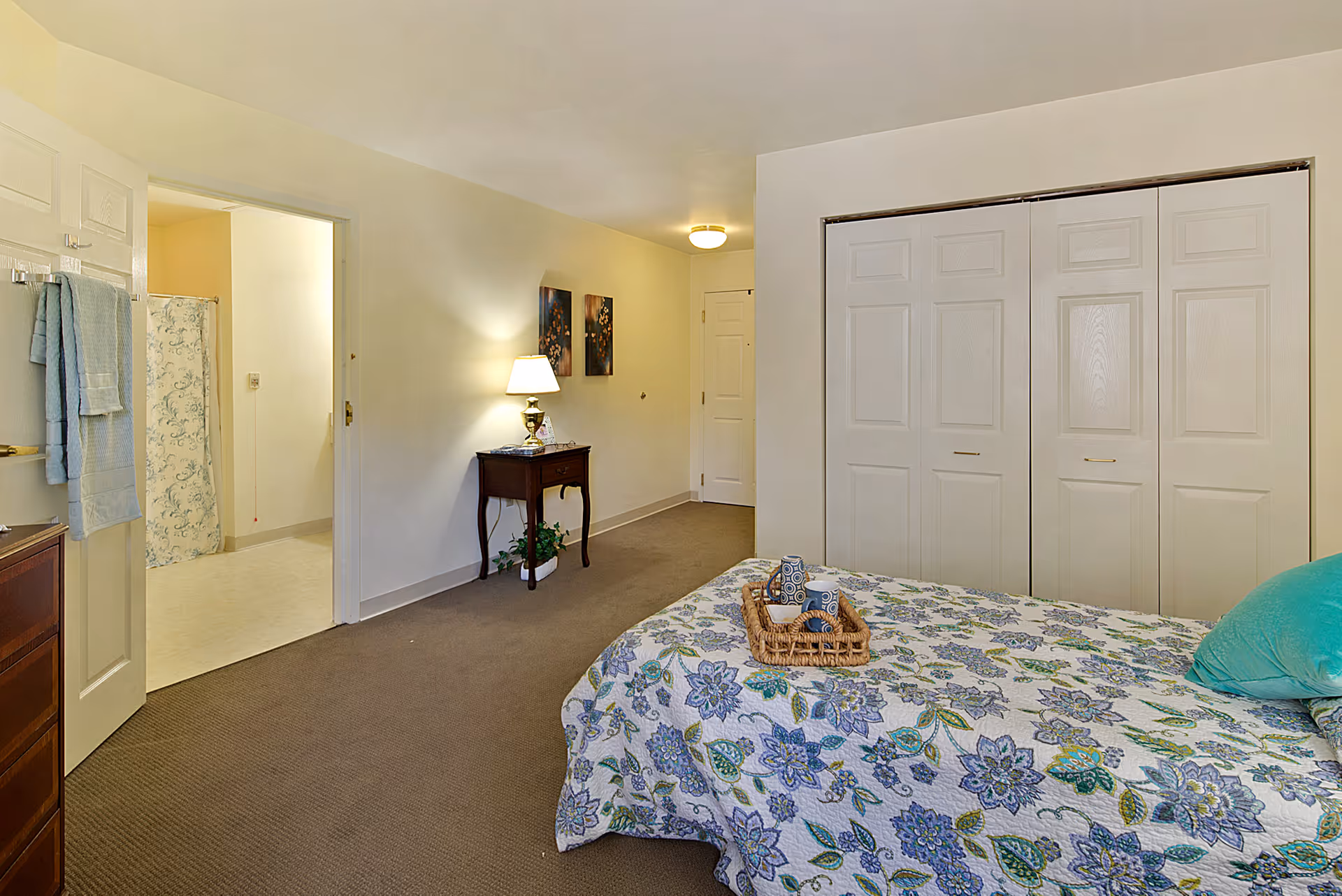 Bright furnished bedroom with a floral quilted bed, large double closets, a nightstand with a lamp, and a doorway leading to a bathroom.