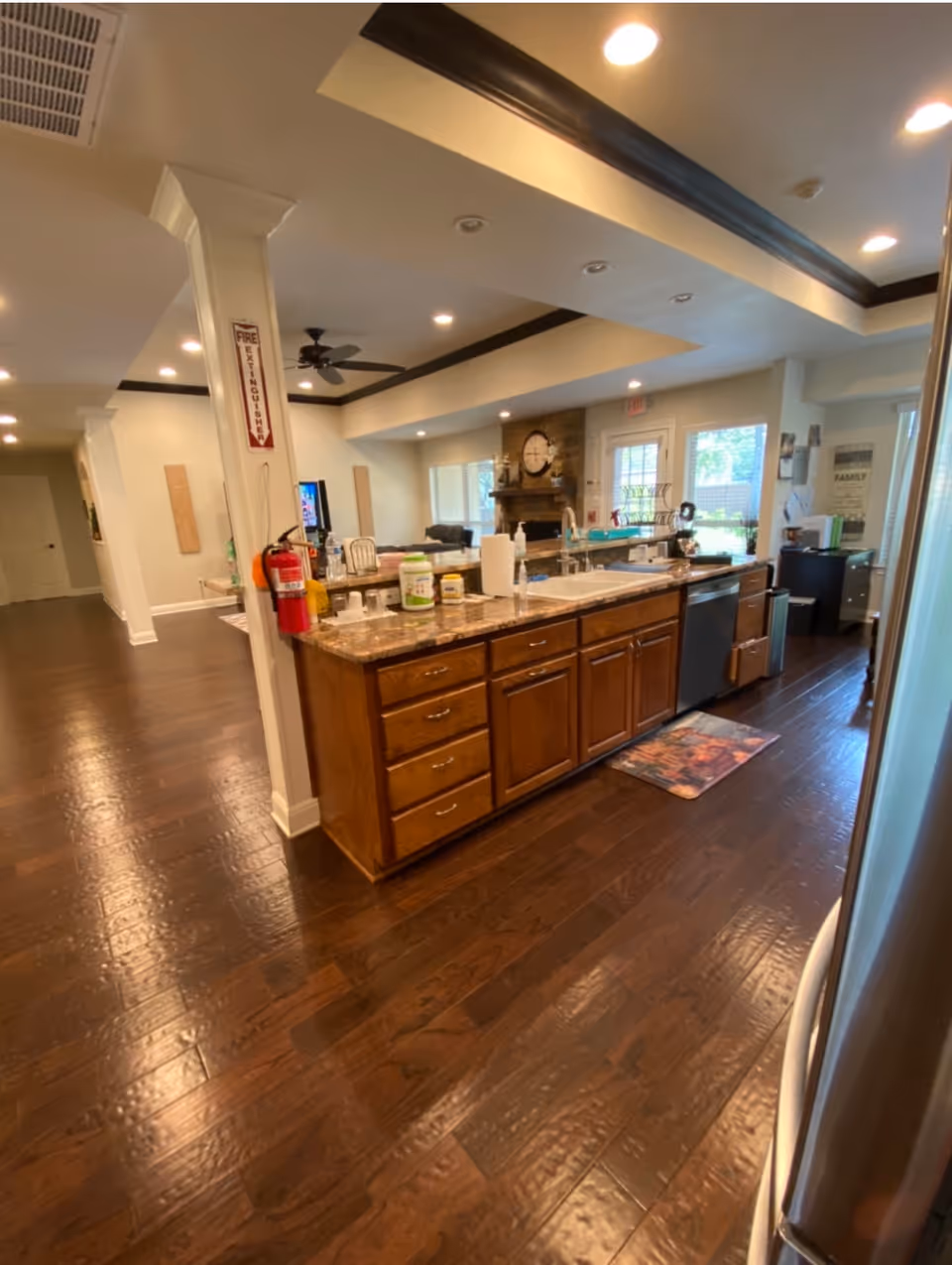 Interior view of a modern assisted living facility kitchen area with dark wooden floors, a large island with granite countertop, sink, and various kitchen items. The background shows a living room area with a fireplace, ceiling fan, and large windows letting in natural light.