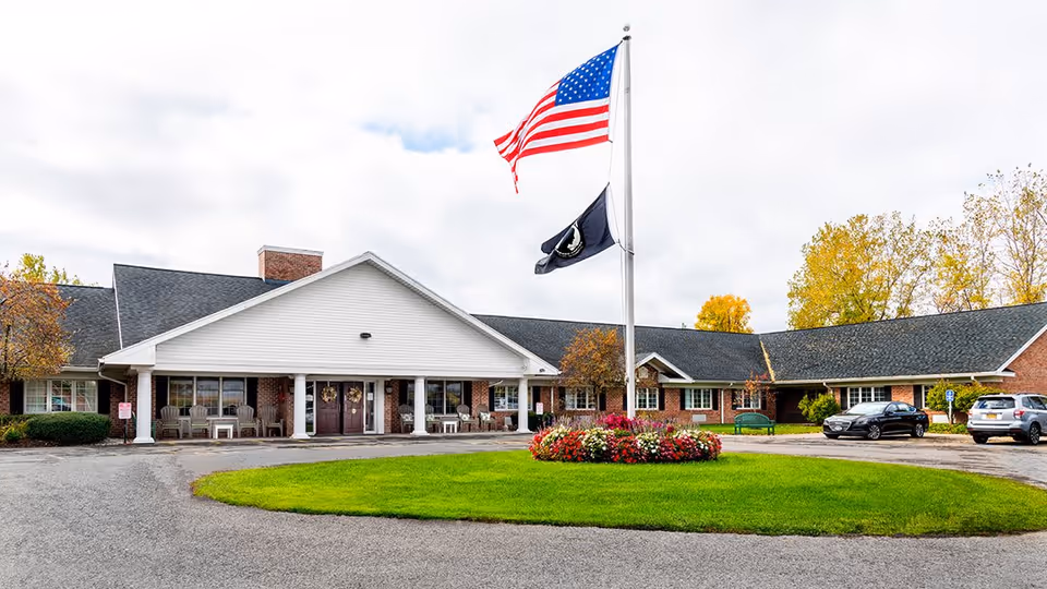 Front exterior view of Elderwood Village at Fairport, a single-story brick building with a white entrance porch supported by columns. Two flags, one American and one black, are flying on a flagpole in front of the building, surrounded by a circular flower bed with colorful flowers. Several cars are parked near the entrance, and trees with autumn foliage are visible in the background under a cloudy sky.