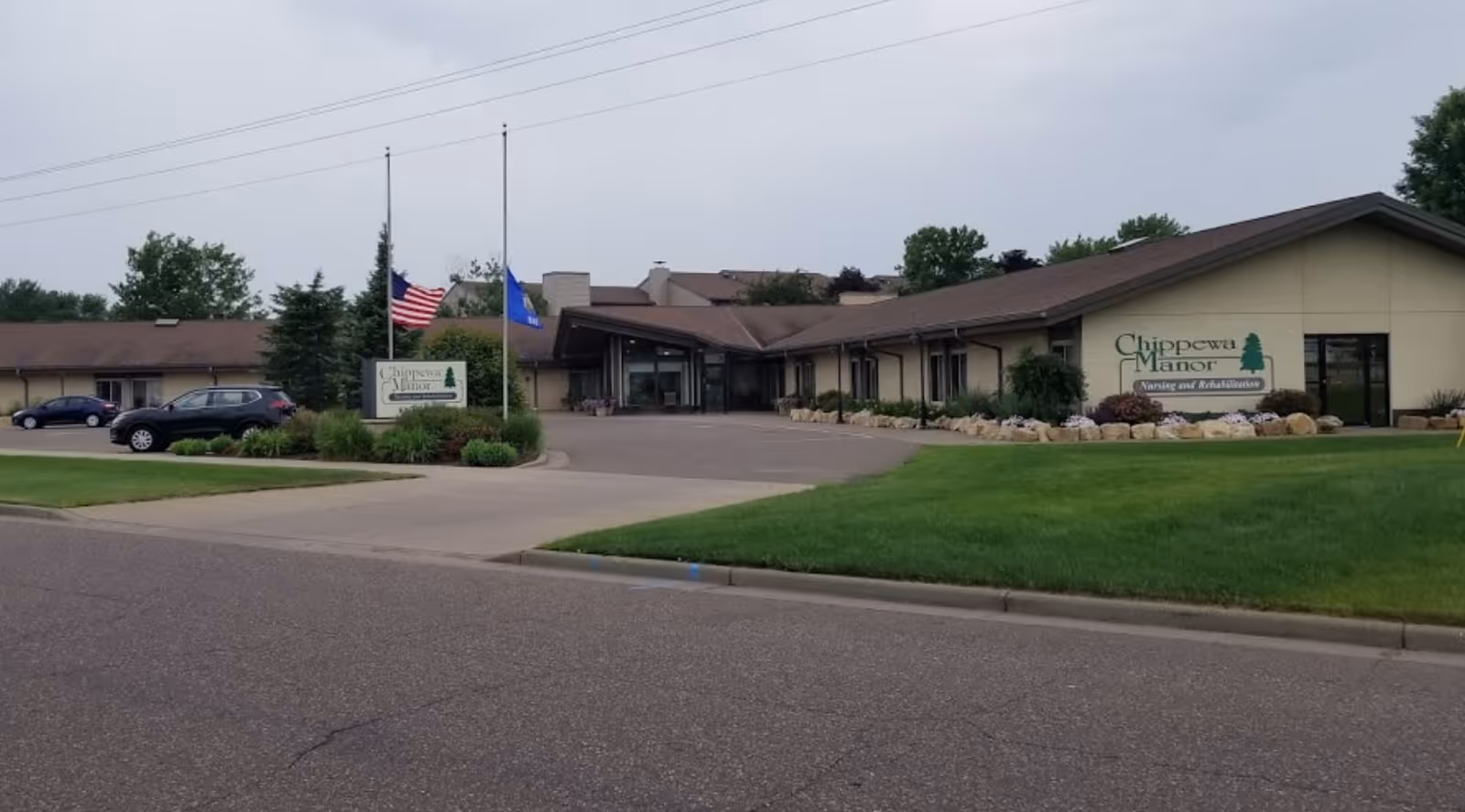 Exterior view of Chippewa Manor nursing and rehabilitation facility with a driveway, green lawn, and two flags at half-mast in front of the building.