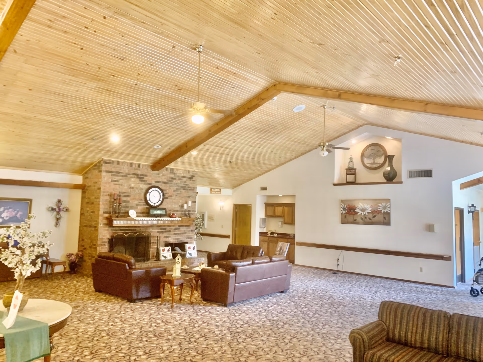Spacious senior living common room with a vaulted wood-paneled ceiling, brick fireplace, leather sofas and decorative accents.