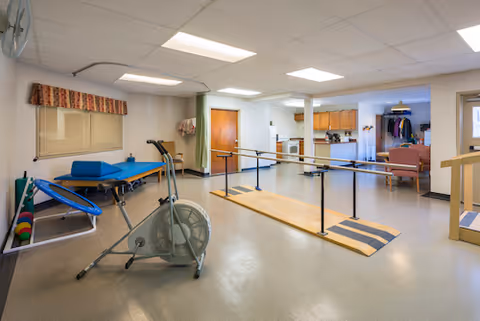 A rehabilitation room featuring parallel bars for walking exercises, an exercise bike, a padded therapy table, and various exercise equipment. The room has a linoleum floor, fluorescent ceiling lights, and a window with a patterned valance. In the background, there is a kitchenette area and a seating area with chairs and a coat rack.