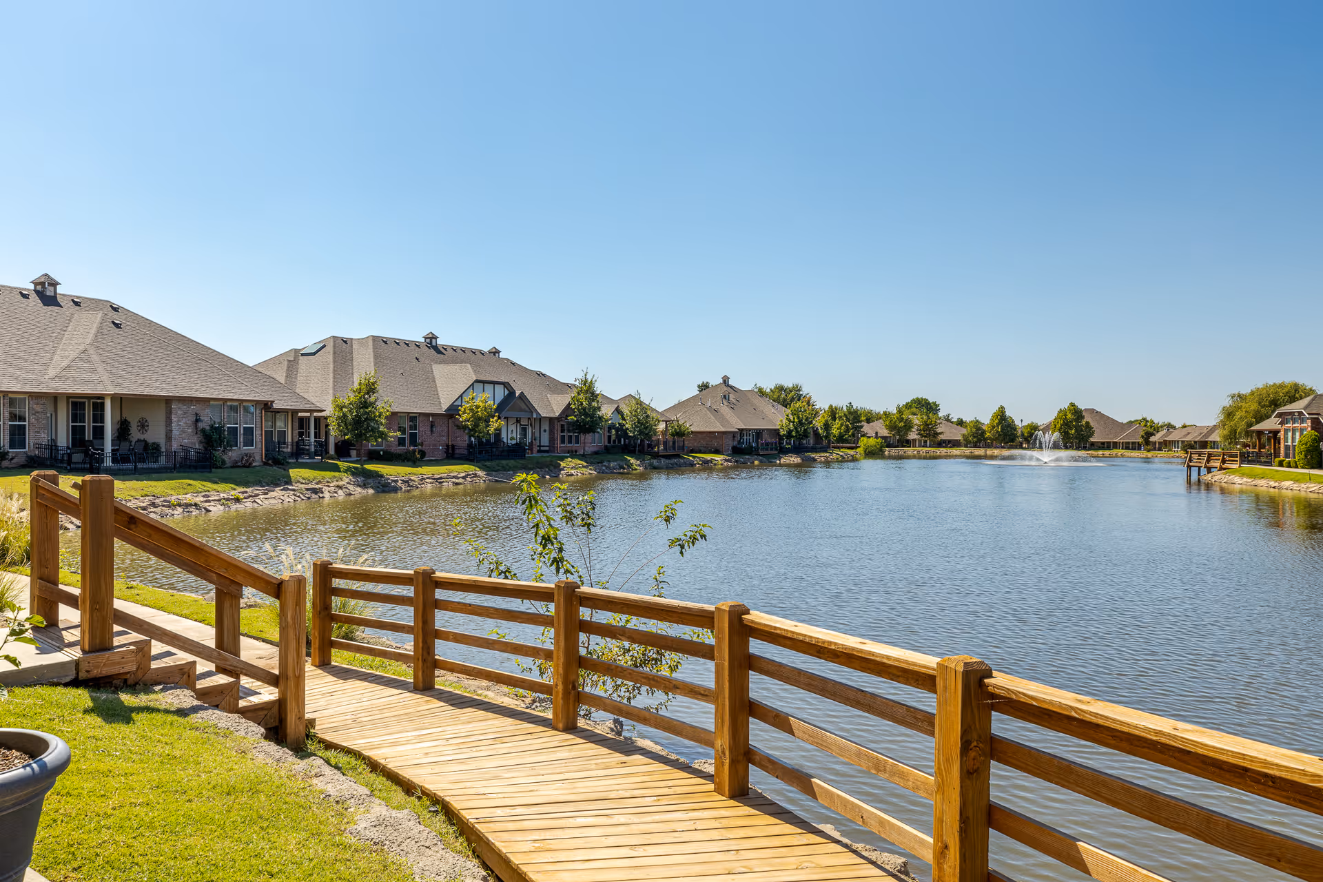 Wooden boardwalk and railing along a pond with houses and a fountain in the distance under a clear blue sky.
