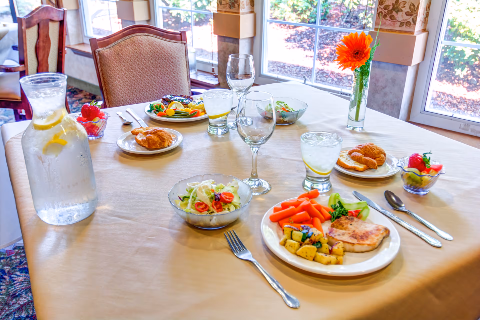 A dining table set with plates of food including grilled chicken with vegetables, salad bowls, croissants, and fruit cups. There are glasses of water with lemon, empty wine glasses, and a pitcher of water with lemon on the table. A single orange flower in a vase is placed near the window with natural light coming in.