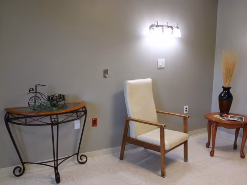 A simple sitting area with a beige upholstered armchair between two small decorative tables, a wall sconce, and a vase.
