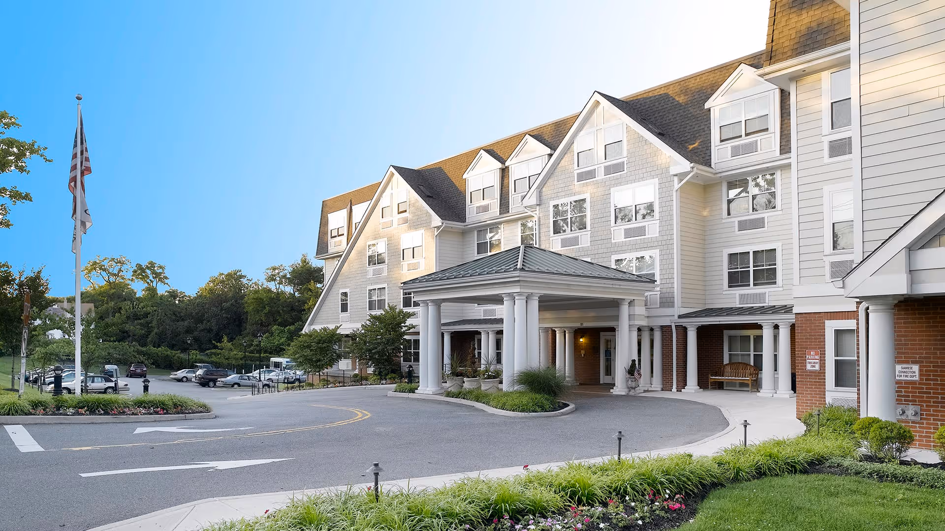 Exterior view of a multi-story senior living facility with a covered entrance supported by white columns. The building has multiple windows and a combination of light-colored siding and brick. There is a driveway in front with landscaped greenery and an American flag on a flagpole to the left.