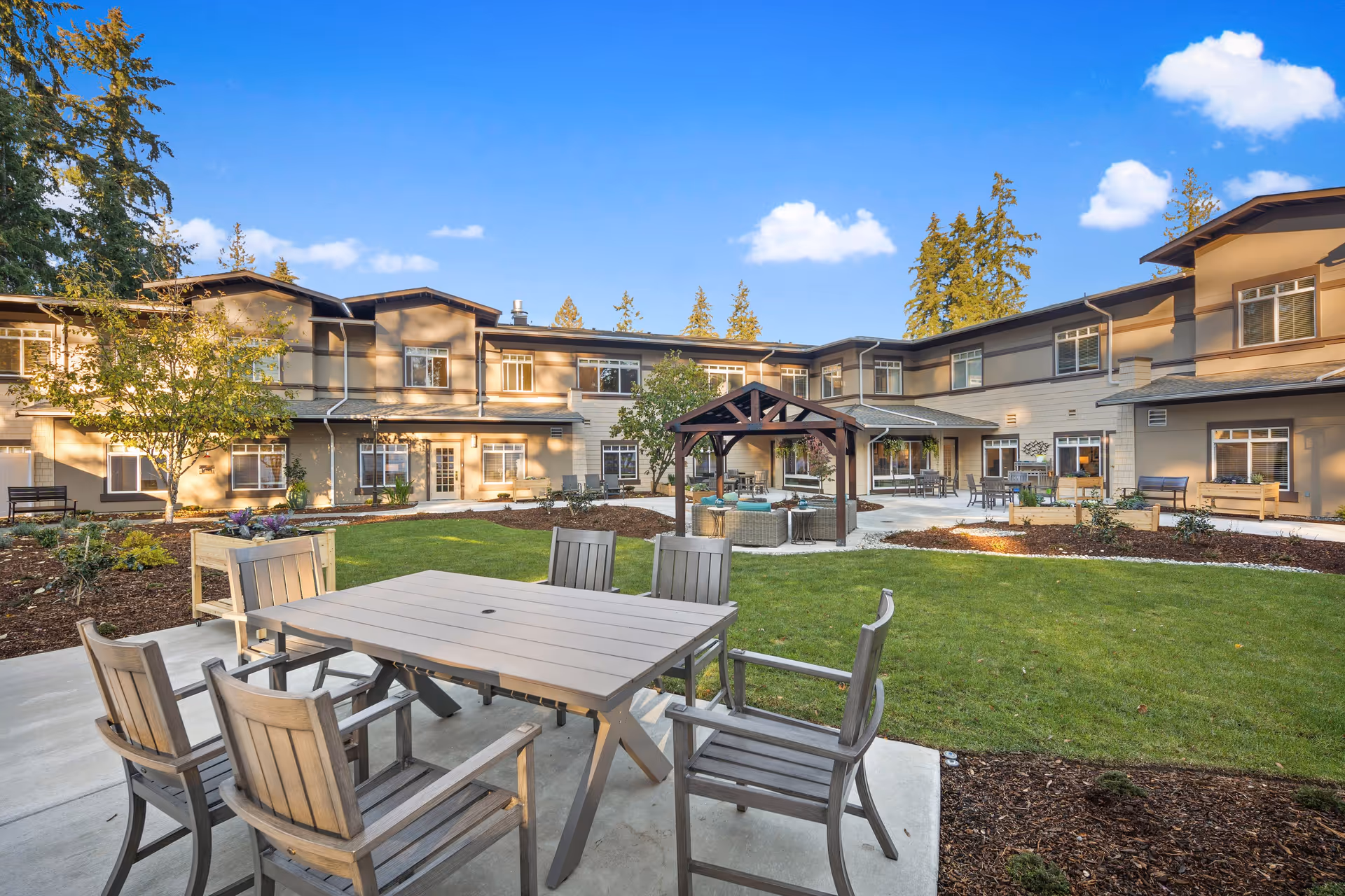 Outdoor courtyard area of Sunrise of Redmond facility with wooden tables and chairs on a concrete patio, green lawn, garden beds, and a wooden gazebo seating area. The two-story building surrounds the courtyard under a clear blue sky with a few clouds.