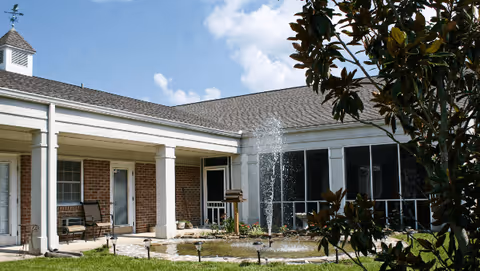 Outdoor courtyard area of a senior living facility with a central water fountain, surrounded by a building with white columns and brick walls, patio chairs, and greenery including a tree in the foreground under a partly cloudy sky.