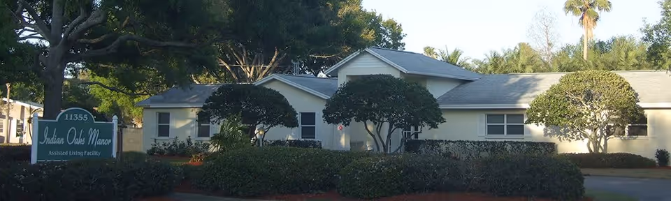 Exterior view of Indian Oaks Manor assisted living facility showing a single-story building with a gray roof surrounded by trimmed bushes and trees under a clear sky.