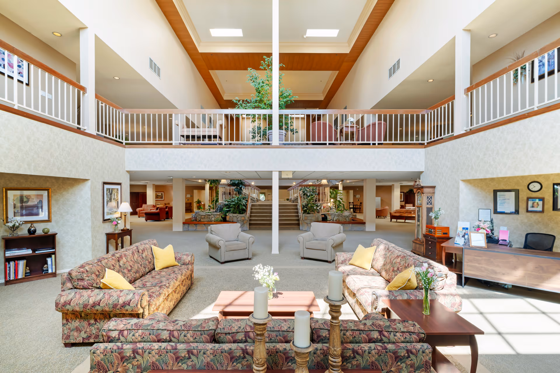 Spacious senior living facility common area with floral patterned sofas, yellow pillows, armchairs, wooden tables with candles and flowers, a reception desk, and a staircase leading to an upper balcony with additional seating and plants under a high ceiling with skylights.