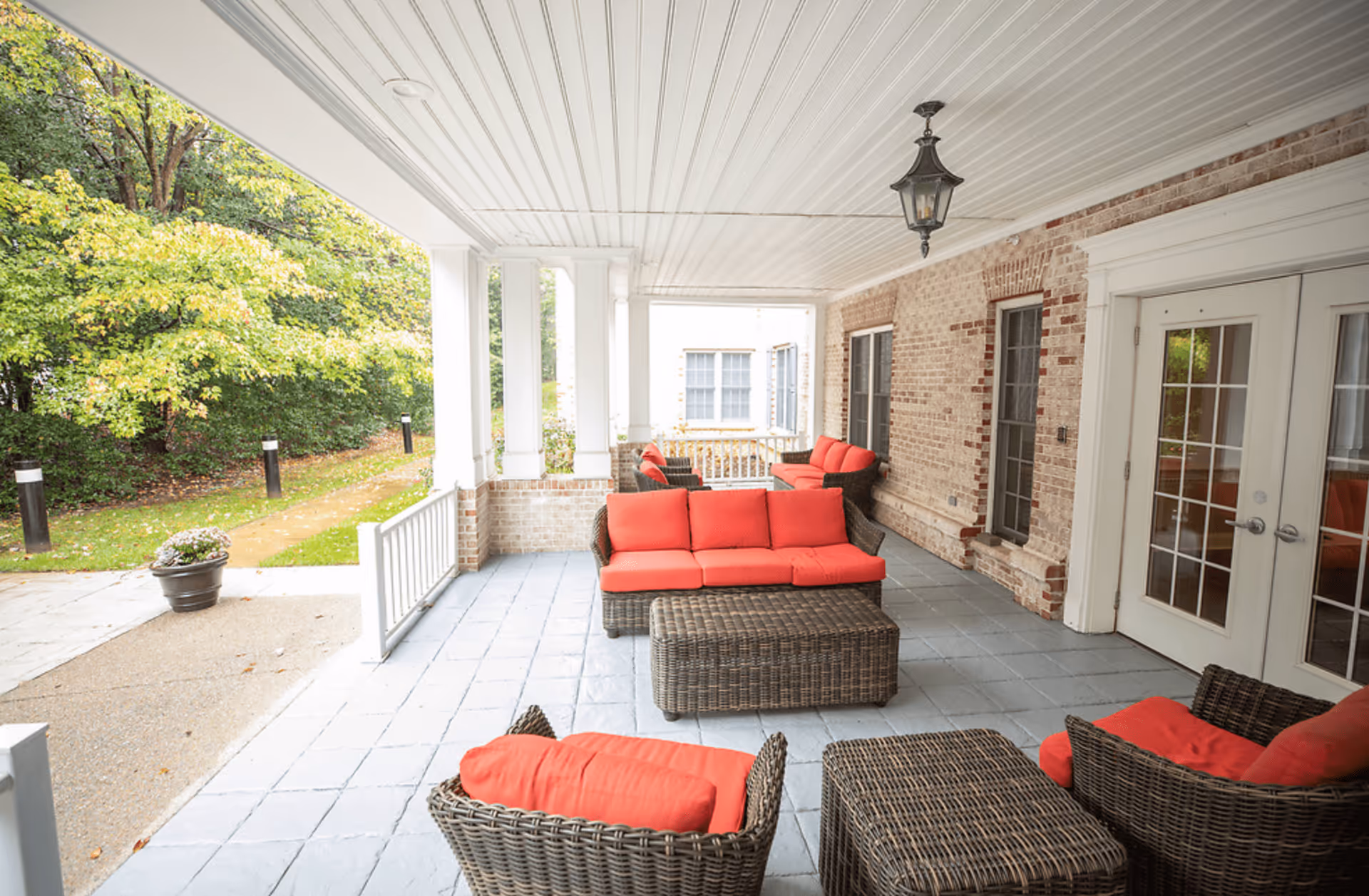Covered outdoor patio area with wicker furniture featuring red cushions, including sofas and chairs, set on a tiled floor. The patio is attached to a brick building with white-framed glass doors and windows. There is a hanging lantern light fixture on the ceiling and greenery visible outside the patio.