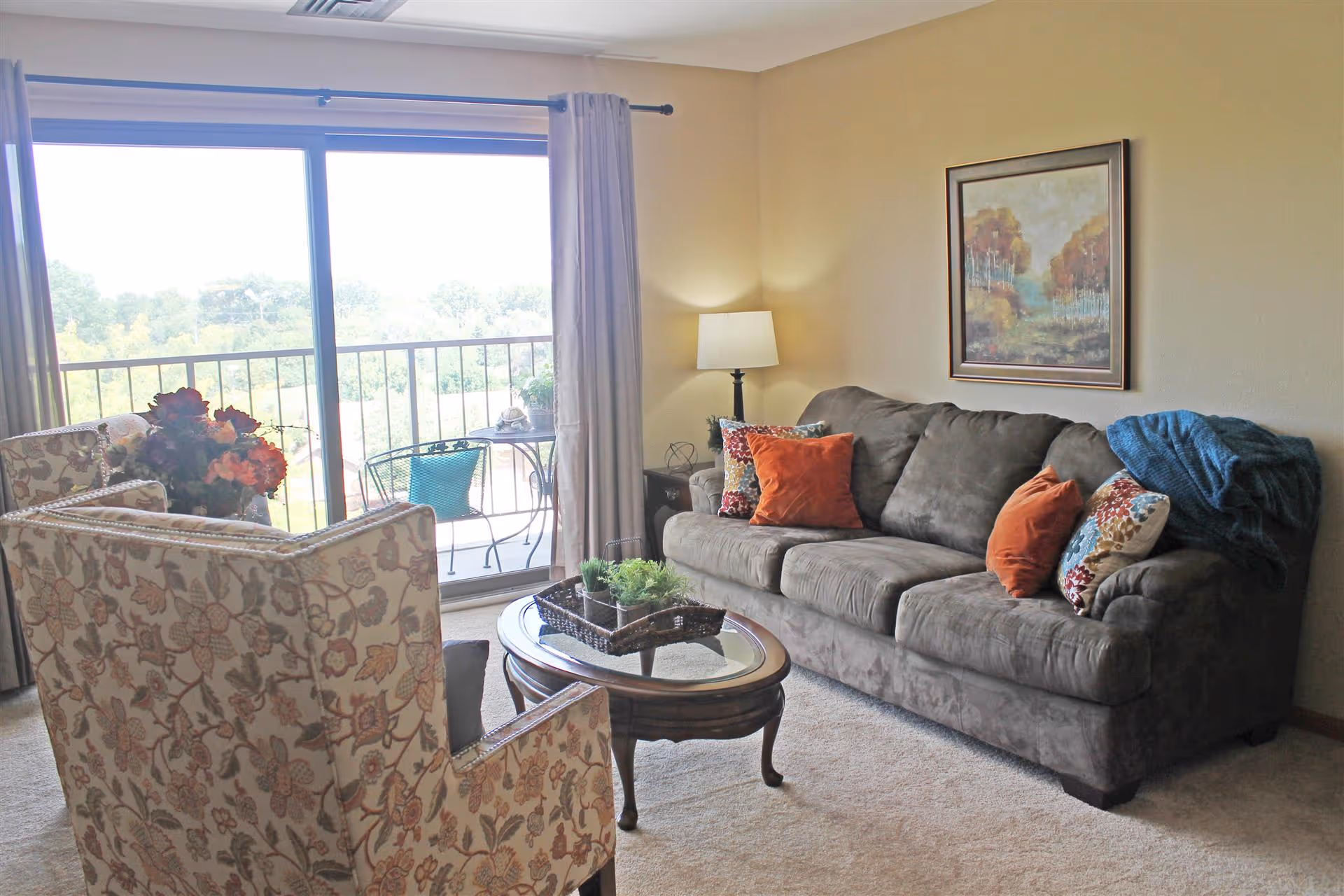 A cozy living room with a gray sofa adorned with colorful throw pillows and a blue blanket. There is a floral patterned armchair, a wooden coffee table with a glass top and a decorative tray with plants. A floor lamp stands in the corner, and a framed painting hangs on the beige wall. Large sliding glass doors lead to a balcony with outdoor seating and a view of trees and greenery.
