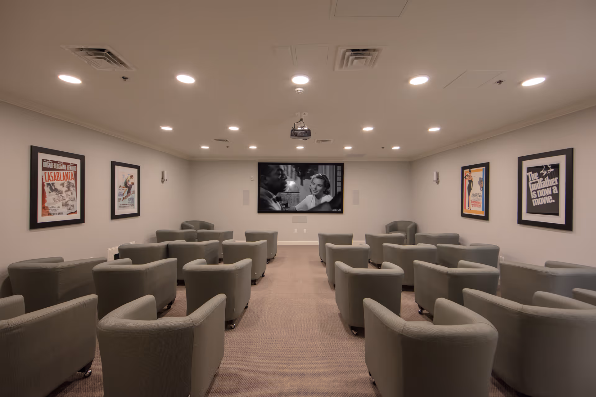 A small movie theater room with multiple rows of gray armchairs facing a large screen displaying a black and white movie scene. The walls are decorated with framed classic movie posters including Casablanca and The Godfather. The ceiling has recessed lighting and a projector is mounted in the center.