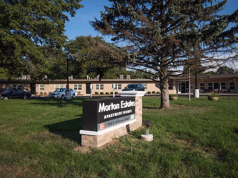 Outdoor view of Morton Estates Apartment Homes with a sign displaying the name and address 935 E Jefferson Street. The building is a single-story structure with several windows, surrounded by grass, trees, and parked cars under a clear blue sky.