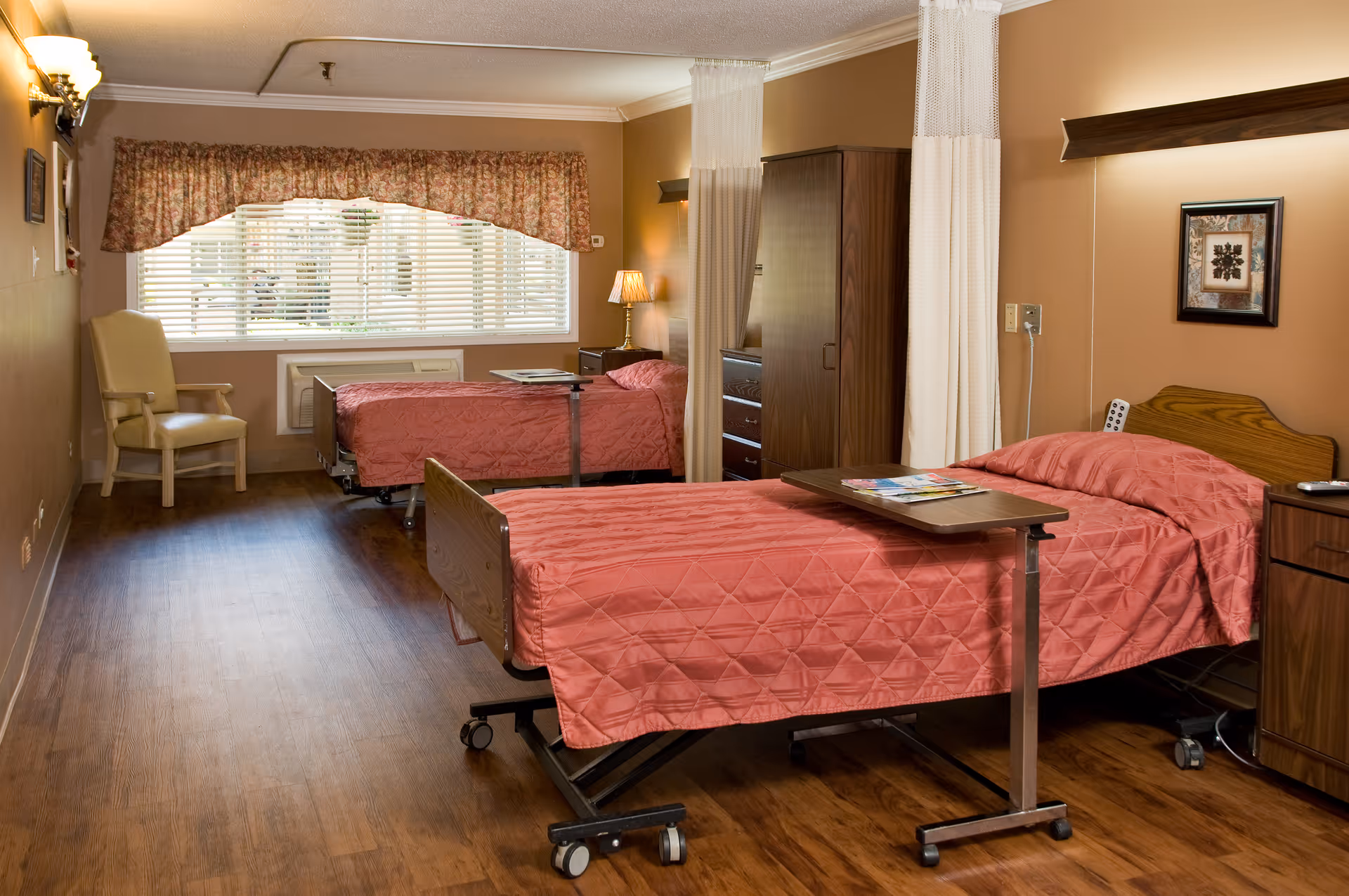 Shared senior living bedroom with two beds covered in pink bedspreads, bedside tables, a chair, and a large window with blinds.