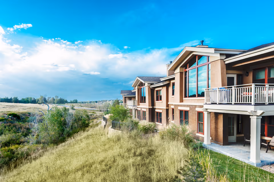 Exterior view of a senior living facility building with large windows and balconies, surrounded by grassy fields and trees under a bright blue sky with some clouds.
